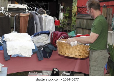 Male Holding A Basket With Linens Inside At A Yard Sale In Front Of A Table With A Sleeping Bag And Clothing Items.