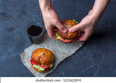 Male Holding Freshly Prepared Grilled Beef Burger Above A Dark Background. Man Serves Burger To The Table. American Snack Close Up.