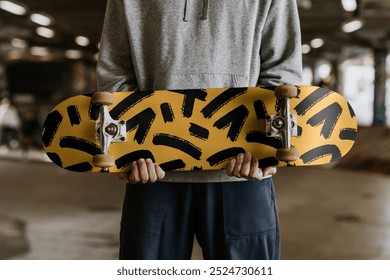 Male holding skateboard with abstract yellow and black design. Skateboard held by male in casual attire. Skateboard with unique yellow and black pattern. Young man in streetwear holding skateboard - Powered by Shutterstock