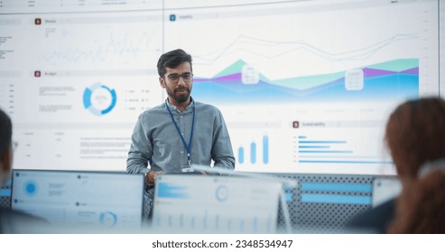 Male Indian Computer Scientist With Tablet Giving Presentation to Diverse Team Of Data Analysts In Front Of Big Digital Screen In Monitoring Room. Colleagues Listening To Training On Automation. - Powered by Shutterstock