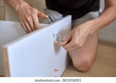 Male individual assembles a drawer using metal fasteners and wooden dowels, kneeling on the floor in a home interior setting