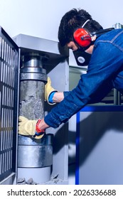 Male Industrial Worker With Protective Mask And Hearing Protectors Removing A Concrete Cylinder From A Testing Machine