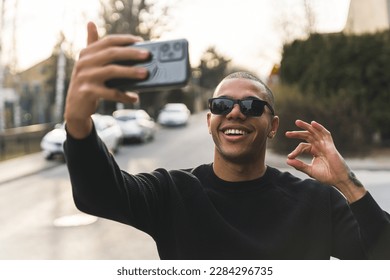 Male influencer doing a stream via smartphone and sharing his favourite outdoor location. Smiling young adult man in sunglasses showing ok sign to viewers. High quality photo