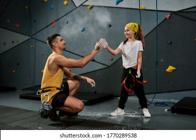 Male instructor teaching little girl, giving high five, applying white dust of magnesia, chalk on hands before climbing wall in bouldering center. Concept of sport and rock climbing. Horizontal shot - Powered by Shutterstock