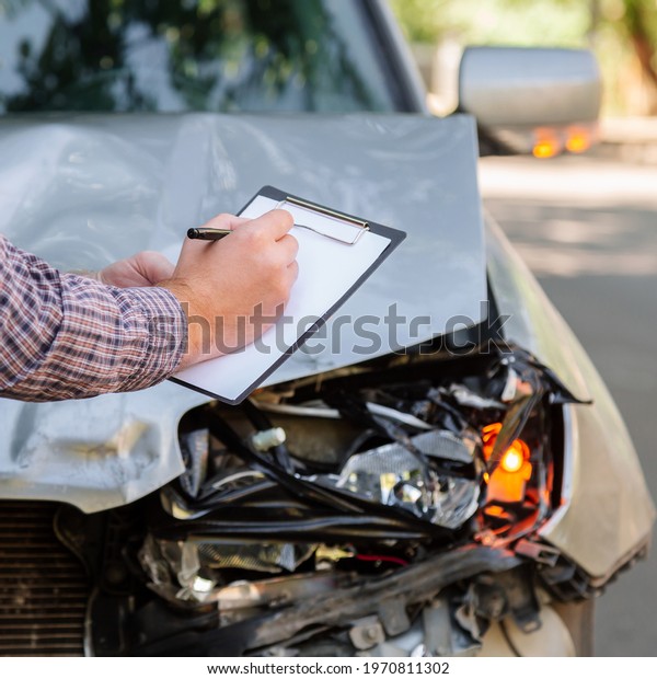 Male insurance agent hands with auto insurance\
blank against Destroyed car in car crash traffic accident on road.\
Smashed broken front auto headlight on car accident. Auto life\
health insurance.