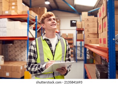 Male Intern Working Inside Warehouse Checking Stock On Shelves Using Clipboard