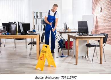 Male Janitor Cleaning Floor With Caution Wet Floor Sign In Office