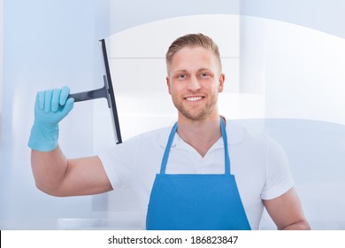 Male Janitor Using A Squeegee To Clean A Window In An Office Wearing An Apron And Gloves As He Works