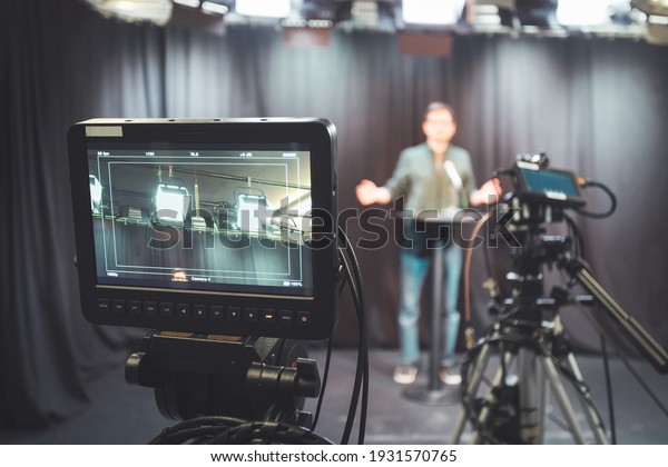Male journalist in a television studio talks into\
a microphone, film cameras