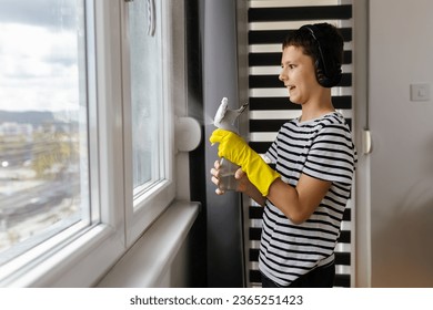 Male kid cleaning the window with the spray and wearing the headphones while listening to the music - Shutterstock ID 2365251423