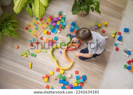 Male kid playing rainbow Montessori wooden natural material early development plaything sitting on floor at home. Cute baby child building labyrinth with colored curved arch ecology cubes blocks