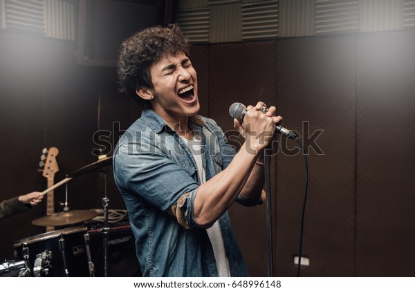 Male lead vocalist singing in studio with\
music instrument\
background.