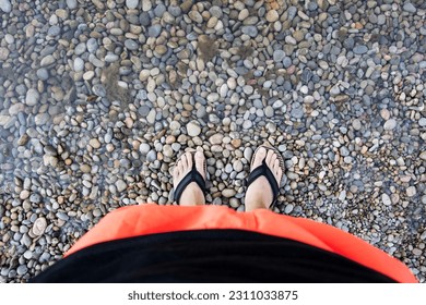 Male legs, orange shorts, swimming trunks, black flip flops on feet, pebble beach and sea water misting feet. Copy space. - Shutterstock ID 2311033875