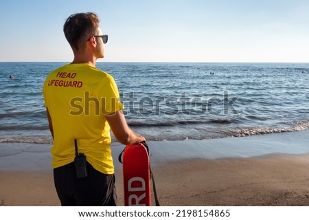 A male lifeguard on the Mediterranean beach watching people in water. Safety while swimming, rear view from handsome brunette male lifeguard on the beach, copy space.