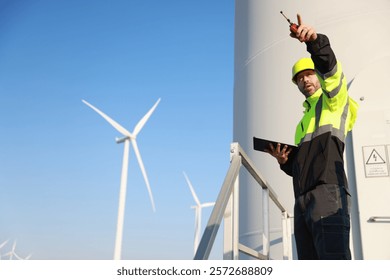 A male maintenance technician in uniform uses a talkie and a digital tablet to work on a station wind turbine. Carbon neutrality and clean energy.