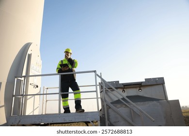 A male maintenance technician in uniform uses a talkie and a digital tablet to work on a station wind turbine. Carbon neutrality and clean energy.