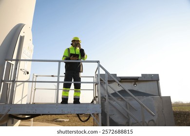 A male maintenance technician in uniform uses a talkie and a digital tablet to work on a station wind turbine. Carbon neutrality and clean energy.
