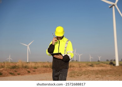 A male maintenance technician in uniform uses a talkie and a digital tablet to work on a station wind turbine. Carbon neutrality and clean energy.