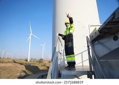 A male maintenance technician in uniform uses a talkie and a digital tablet to work on a station wind turbine. Carbon neutrality and clean energy.