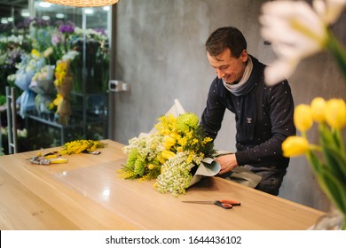 Male Make Spring Bouquet. Man Florist Wrapping Beautiful Bouquet Of Spring Flowers In Pack Paper On The Wooden Table. Beautiful Flower Composition Of Yellow Flowers