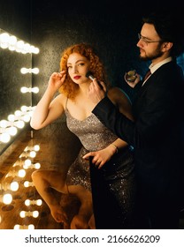 Male Makeup Artist Applying Powder On Female Face. Curly Red Haired Woman In Evening Dress And Handsome Bearded Man In Suit Near Big Mirror In Dressing Or Make Up Room. Couple Before Going To Party