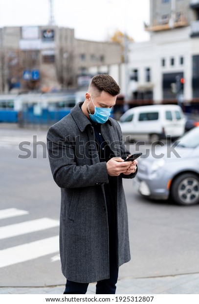 Male in mask types\
message outdoors. Man in suit, coat and protective mask stands in\
the city center.
