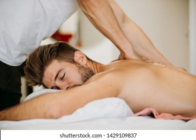 Male to male massage from young Asian therapist giving massage to muscle gym fit Caucasian man who laying on the bed in massage room. Remedial oil massage is performing on client.