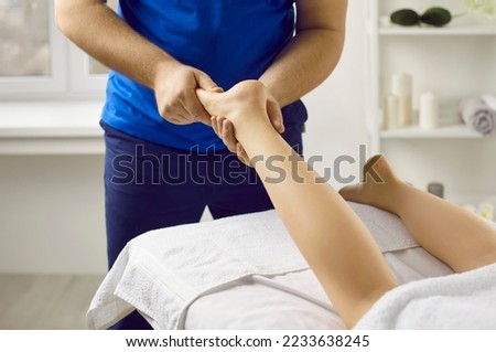 Male masseur in health clinic or massage parlor does hand massage of young woman's feet. Cropped image of woman lying on massage table and receiving acupressure of foot which affects flow of energy.