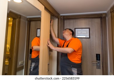 A male master in bright overalls installs a new wardrobe in the hotel. - Shutterstock ID 2256583199