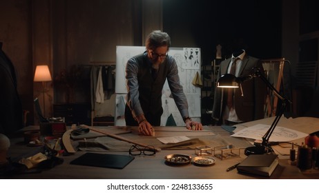 Male mature tailor in luxury designer atelier or tailoring studio. He works with sketch of future garment on table with fabric and tools. Mannequin with tailored suit. Fashion and hand craft concept. - Powered by Shutterstock