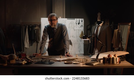 Male mature tailor works in luxury designer atelier or tailoring studio, looks at camera. Table with threads, fabric and tools. Tailored suits and board with sketches on background. Fashion concept. - Powered by Shutterstock