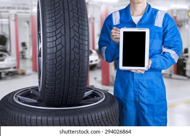 Male Mechanic With Blue Uniform Showing Empty Tablet Screen Near The Tires, Shot At Workshop