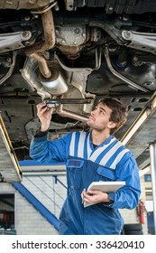 Male Mechanic Examining Exhaust System Of Car With Flashlight At Auto Repair Shop