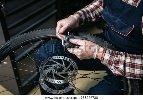 Male mechanic working in bicycle repair shop,\
mechanic repairing bike using special tool, wearing protective\
gloves. Young attractive serviceman fixing customer\'s bicycle wheel\
at his own workshop.