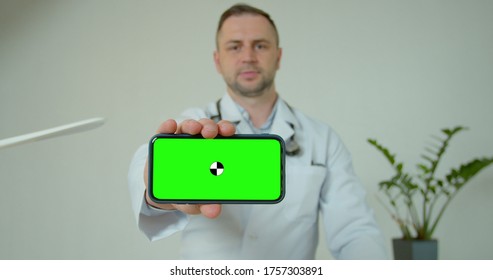 Male Medical Professional Displaying Mobile Smartphone With Greenscreen. Doctor Man Holding Phone With Green Screen Up To Camera. Green Screen On Phone Held By Scientist. Male Doctor Demonstrating