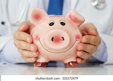 Male medicine doctor wearing blue tie holding and covering happy funny smiling piggybank in hands closeup. Medical service economy, health care savings and insurance concept. Focus on piggy bank