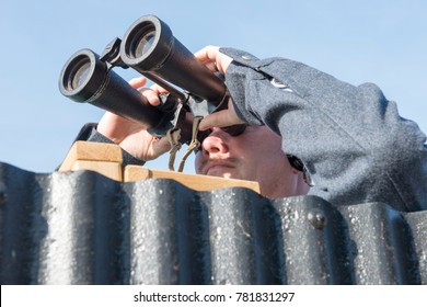 Male military personnel peering through a pair of huge binoculars behind a corrugated iron sheet - Powered by Shutterstock