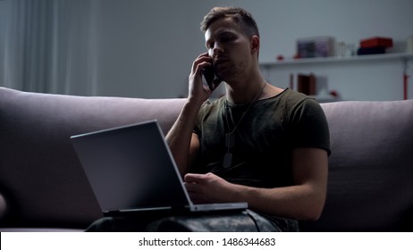 Male Military Spy Sitting With Laptop On Sofa And Talking Smartphone At Night