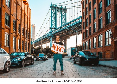 Male Model Holding NYC Board With Manhattan Bridge In Background Between Manhattan And Brooklyn Over East River With Two Brick Buildings On A Sunny Day In Washington Street In Dumbo, Brooklyn, NYC