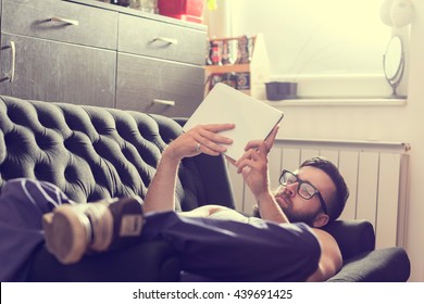 Male Model Lying On A Couch In A Living Room, Surfing The Web On A Tablet Computer