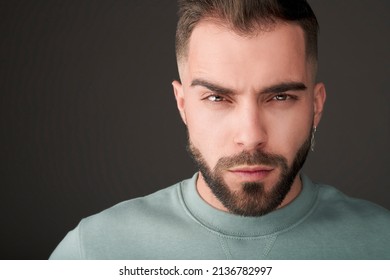 Male Model Shot In Studio. Serious And Confident Expression. Angry Or Upset Look And A Casual Style. Headshot Of Man With A Beard, Brown Hair, Brown Eyes On A Grey Solid Background. 