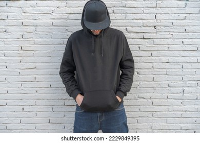 Male model wearing a black sweatshirt and a cropped body, covering his face with a black cap, leaning against a white wall. - Shutterstock ID 2229849395