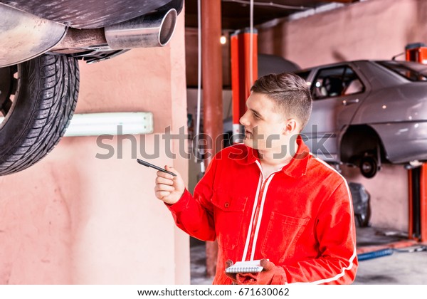 Male motor mechanic standing making notes\
underneath a lifted car near working equipment for repair and\
diagnostic in auto repair\
service