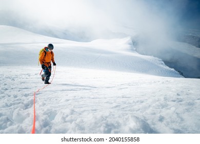 A male mountaineer walking uphill on a glacier with orange jacket. On the way up to the top. Whole route over the glacier. Mountaineering and alpinism on alpine peaks. Mountain look into misty valley