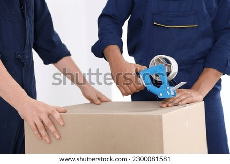 Male movers packing box with adhesive tape indoors, closeup