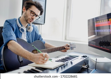 Male music arranger composing song on midi piano and audio equipment in digital recording studio. Man plays guitar and produce electronic soundtrack or track in project at home.