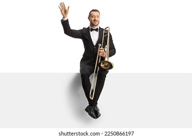 Male musician sitting on a blank panel with a trombone and waving isolated on white background