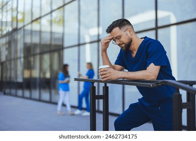 A male nurse in blue scrubs appears troubled while holding a coffee cup outside a medical facility, reflecting the stress of healthcare professionals. - Powered by Shutterstock