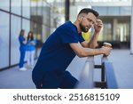 A male nurse in blue scrubs leans on a railing while holding a coffee cup. He appears thoughtful as he takes a break outside a medical facility.