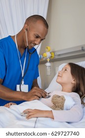 Male Nurse Checking Blood Pressure Of Little Patient Lying On Bed. African Doctor Examining Child Patient At Hospital. Doctor Man With Stethoscope Examining Young Girl In Hospital.